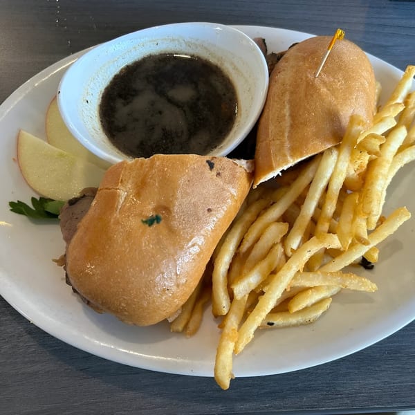 A French dip sandwich with a side of fries, au jus, and apple slices on a white plate.